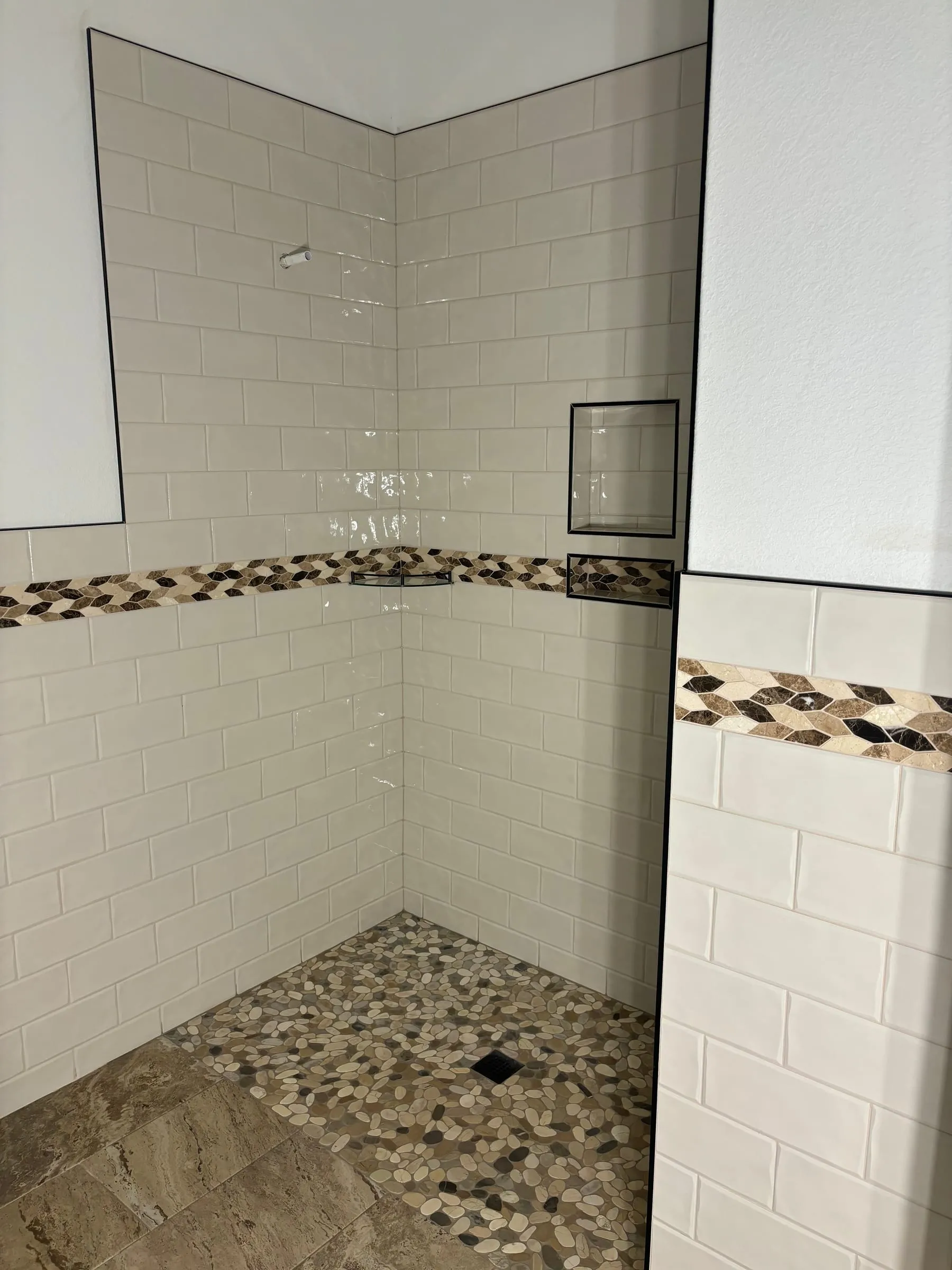 Corner of a subway tile shower with a mosaic accent band and pebble floor