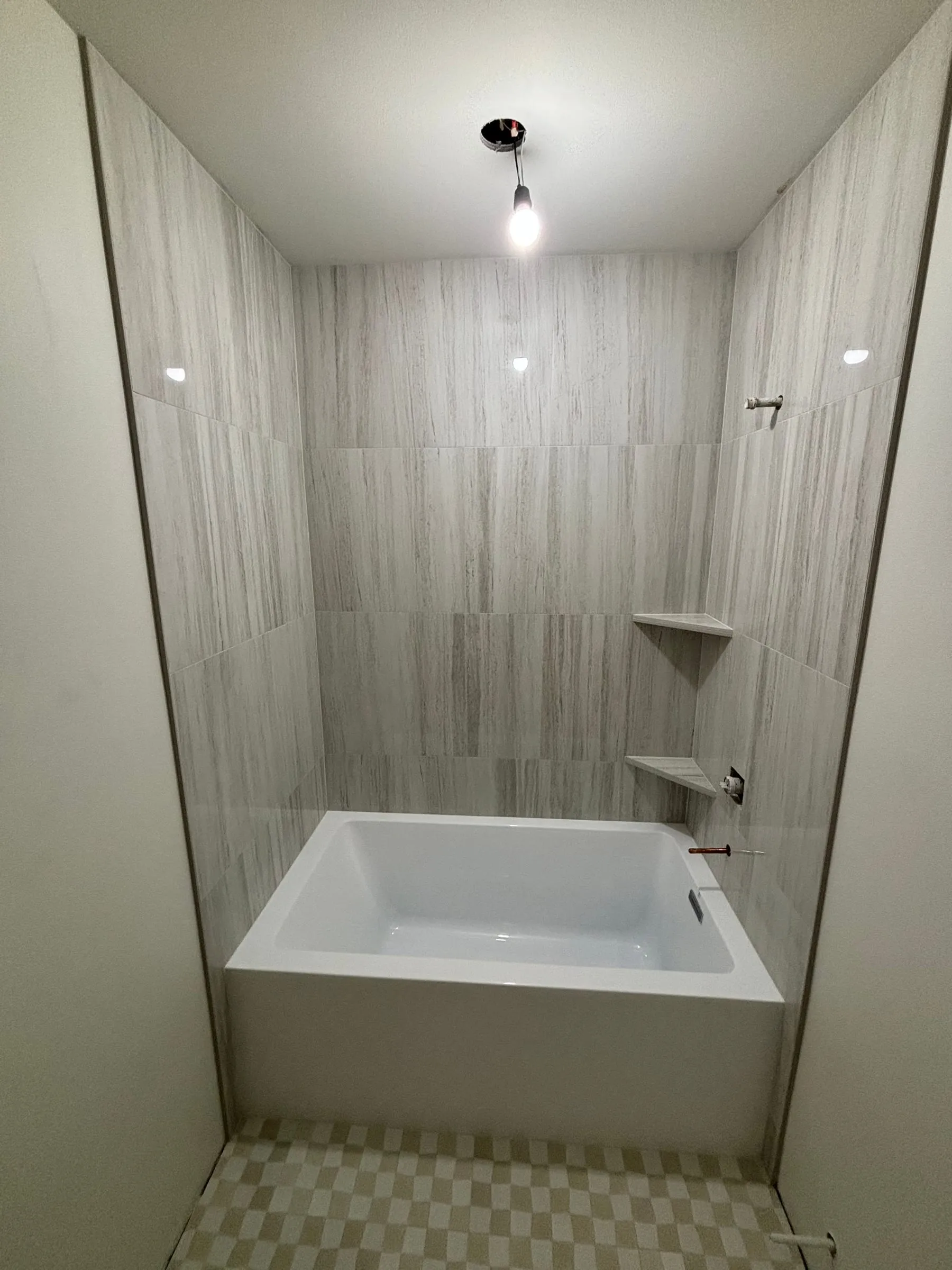 Tub alcove in wood-grain stone-look tile with corner shelves and a checkered floor