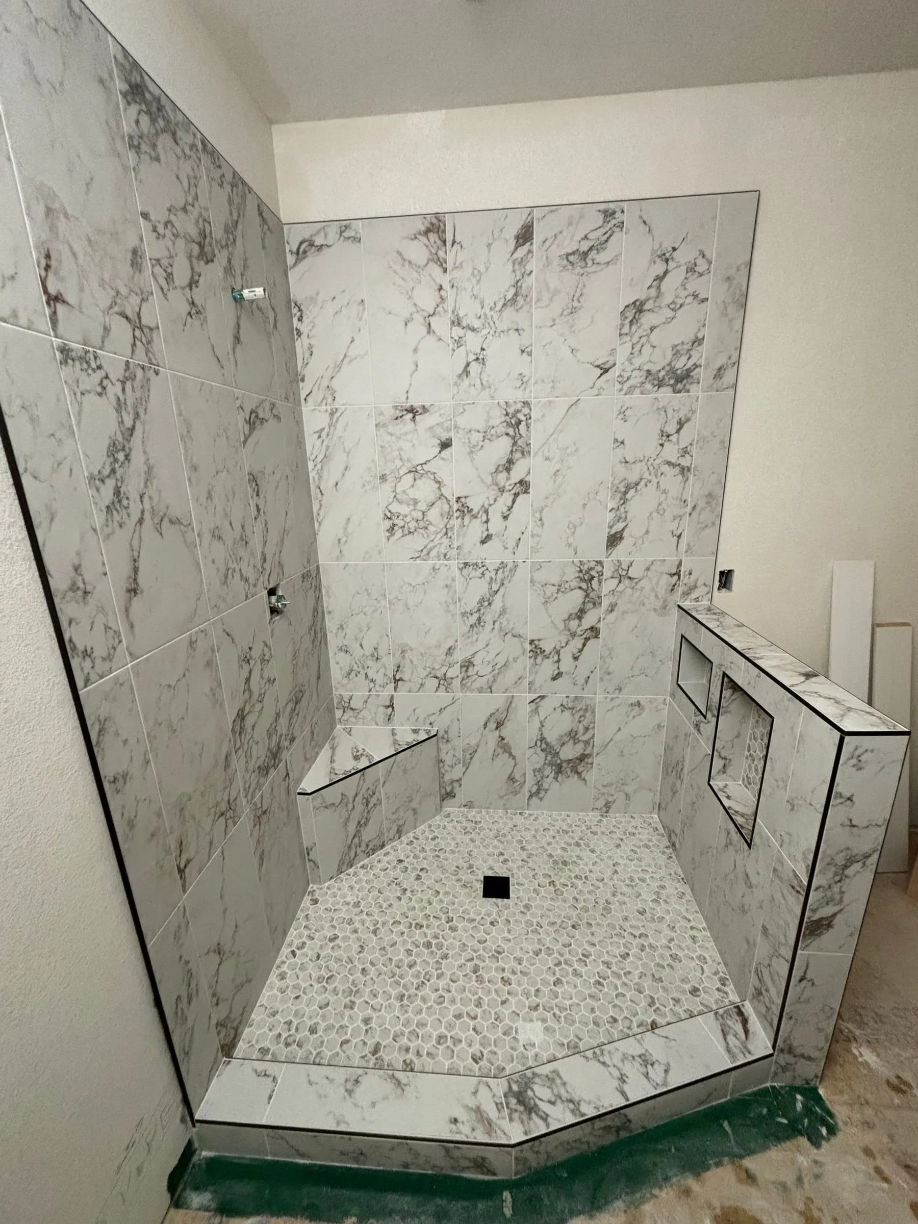 Corner shower in large marble-look tile with a bench, niche, and mosaic floor, during installation