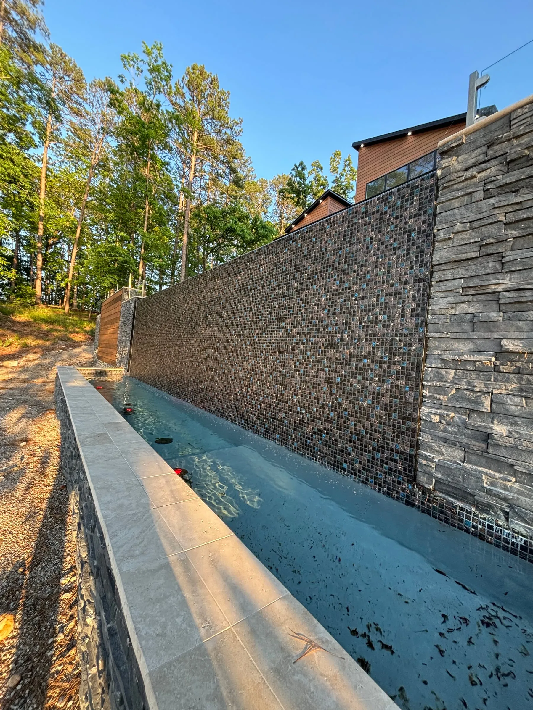 Pool wall faced in glass mosaic tile beside a stacked-stone wall, with coping along the edge