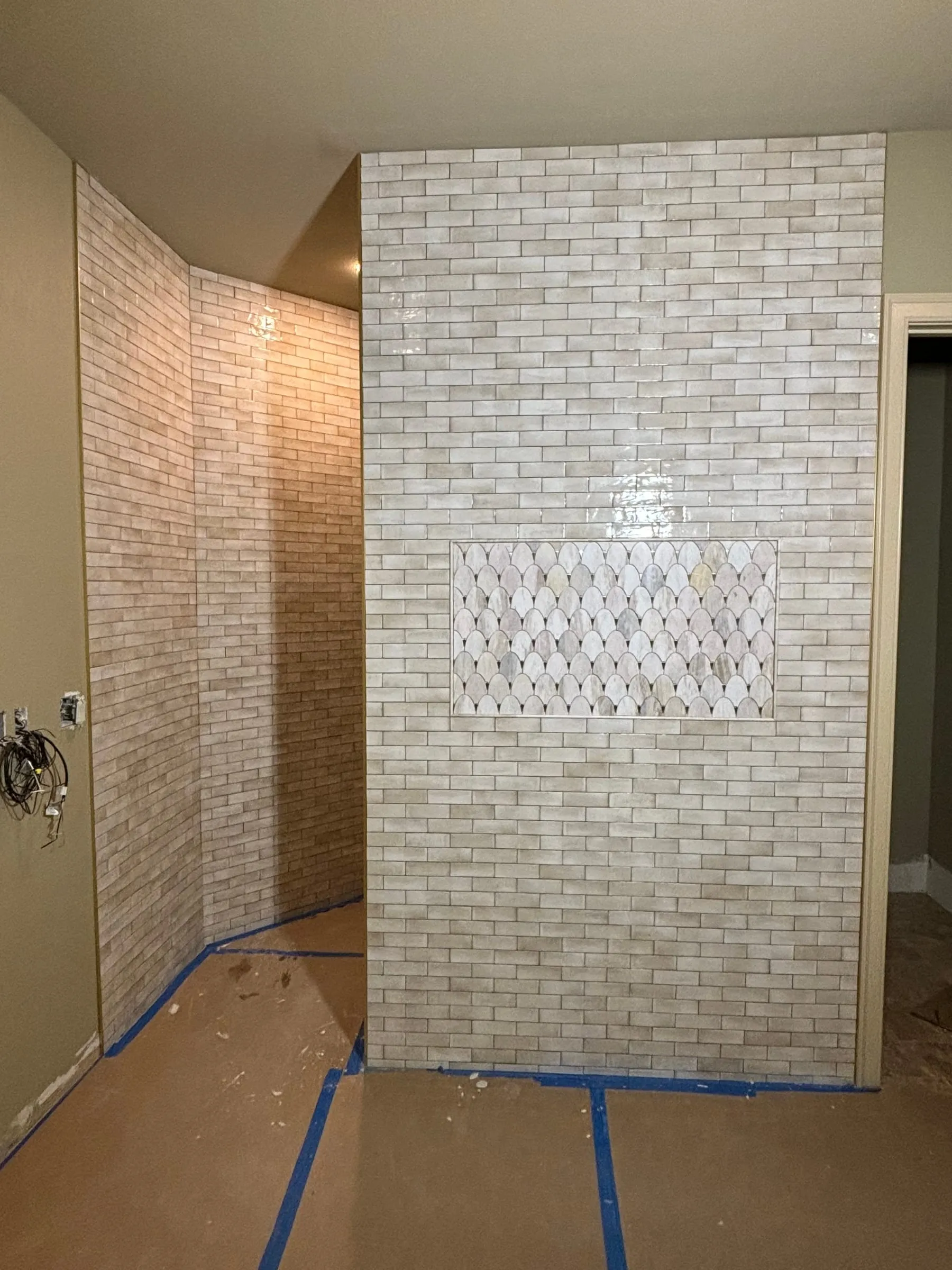 Shower wall in glossy cream brick-look tile with a marble fish-scale mosaic panel, during installation